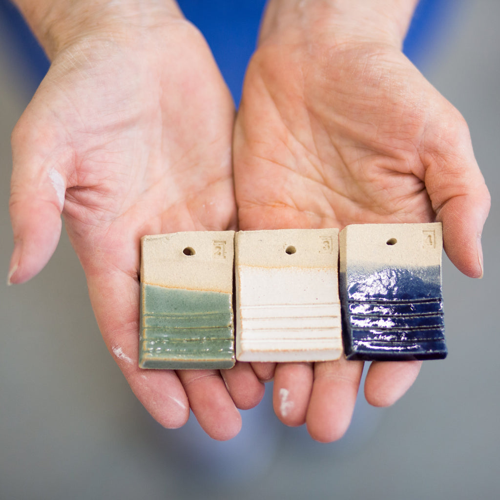Hands holding three small ceramic tiles with different color patterns against a blurred background