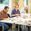 Three people working at a table with sewing machines and materials in a bright room.