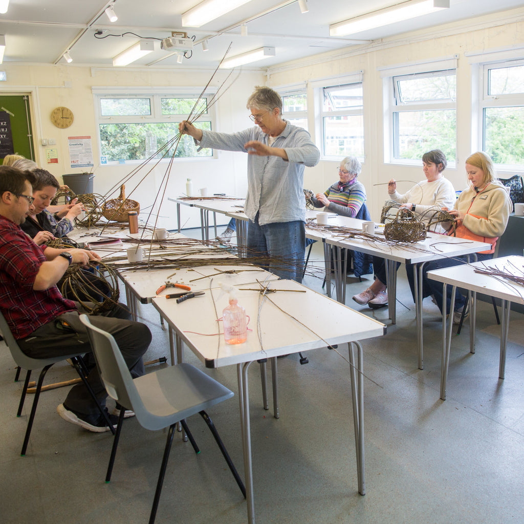 A group of people seated and standing around tables in a bright classroom or workshop, working with long strands of material, likely learning a craft or knitting class.