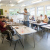 A group of people seated and standing around tables in a bright classroom or workshop, working with long strands of material, likely learning a craft or knitting class.