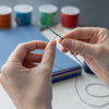 Close-up of a person's hands holding a blue needle and thread over a stack of colorful spools of thread on a white desk.