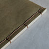 A close-up of a handmade book or journal with a textured olive green cover, bound with visible thread and white paper edges, resting on a light gray surface.