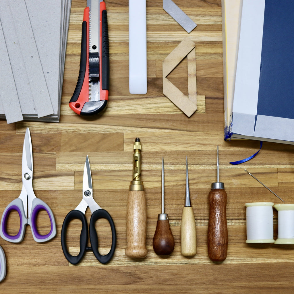 A flat-lay arrangement of various leathercraft and sewing tools on a wooden surface, including scissors, a utility knife, wooden carving tools, spools of thread, and a folded piece of blue material.