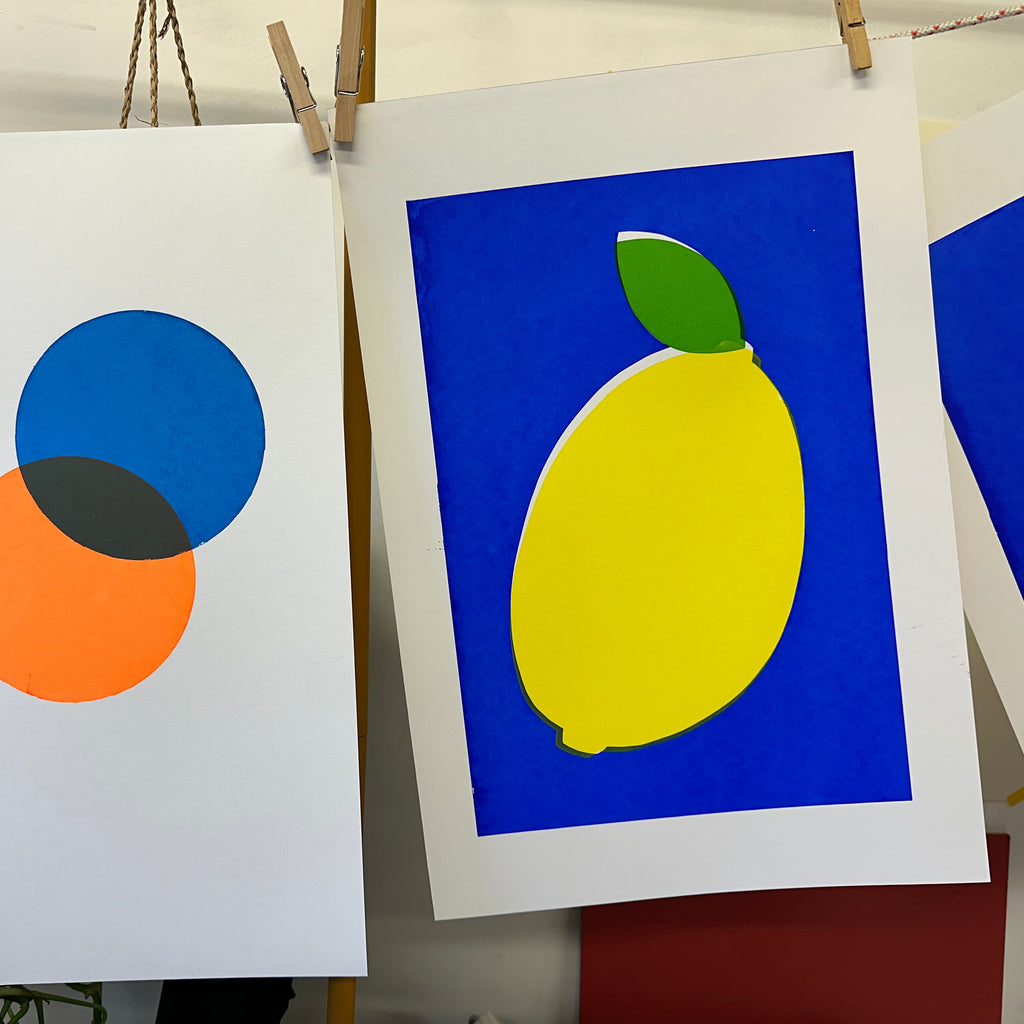 Two prints on paper with abstract designs, one featuring a yellow lemon on blue and the other with geometric shapes.