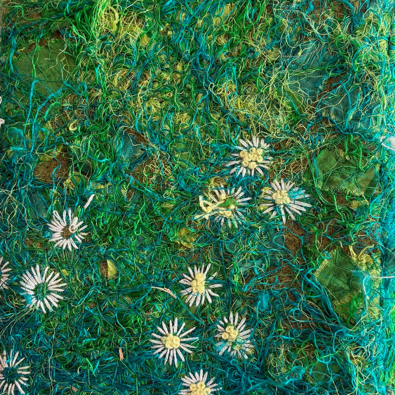 Textile art piece with green and brown hues and white floral patterns on a textured surface.