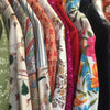 A close-up row of hanging patterned garments or fabric pieces in multiple colors, including cream with illustrated motifs, red, pink glitter trim, and a bright floral print with blue, pink, orange, and green.