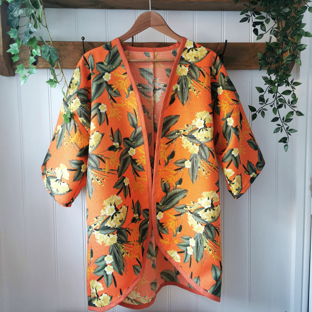An orange kimono-style cardigan with a green and yellow floral print, hanging on a wooden hanger against a white paneled wall with decorative greenery.
