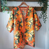 An orange kimono-style cardigan with a green and yellow floral print, hanging on a wooden hanger against a white paneled wall with decorative greenery.