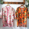 Two lightweight floral open-front cardigans hang on wooden hangers from a wooden coat rack against a white paneled wall. The left cardigan is light pink with red and green floral print and pink trim.