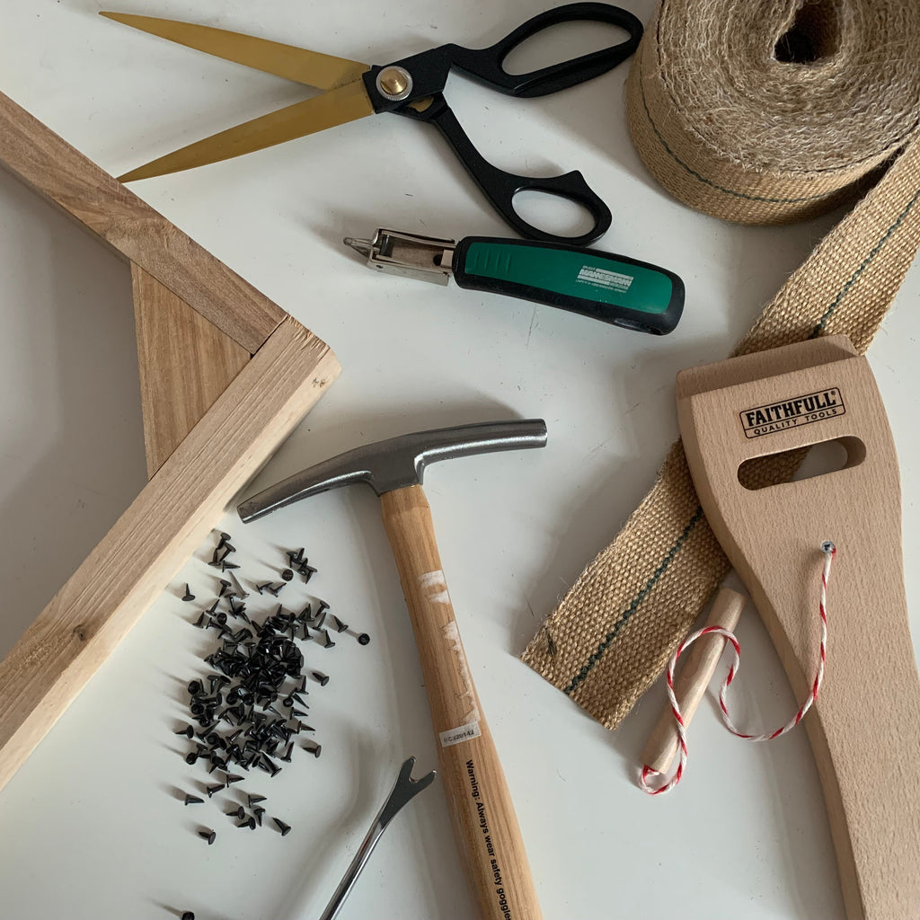 A flat-lay of hand tools and materials for woodworking, including a wooden frame, a claw hammer, a green-handled nail gun, scissors, twine, and a burlap ribbon on a white surface.