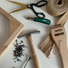 A flat-lay of hand tools and materials for woodworking, including a wooden frame, a claw hammer, a green-handled nail gun, scissors, twine, and a burlap ribbon on a white surface.