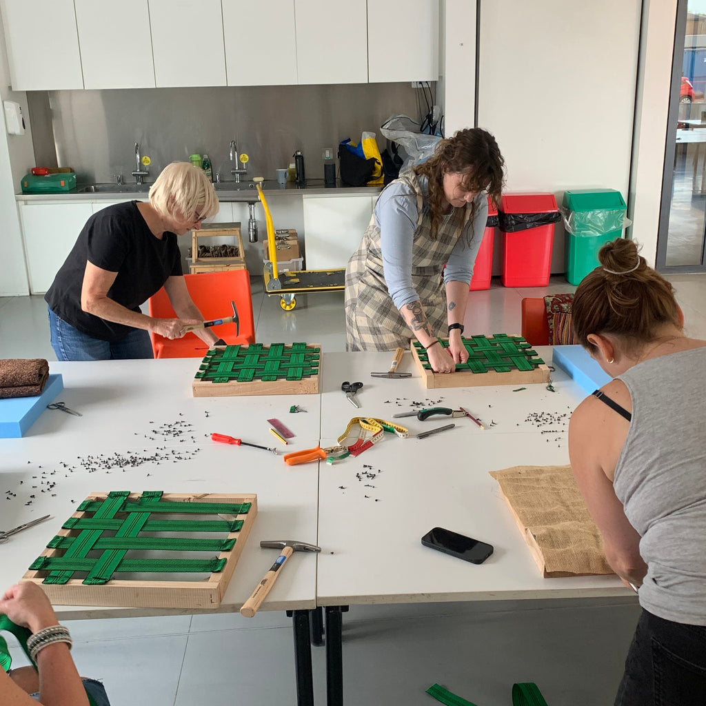 Three people are gathered around a white table in a workshop or classroom setting, assembling green wooden structures using tools and screws. The room has white walls and various workshop items in the background.