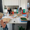 Several people are gathered around a long table in a bright modern room, working on handmade wooden crafts with tools, wood panels, and colorful materials. The scene shows a real environment with multiple people present, so the subject type is Lifestyle.