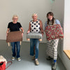 Three women standing in a room, each holding a handmade upholstered furniture or textile item. The items include a brown upholstered stool, a gray upholstered chair, and a red patterned cushion, set against a plain white wall.