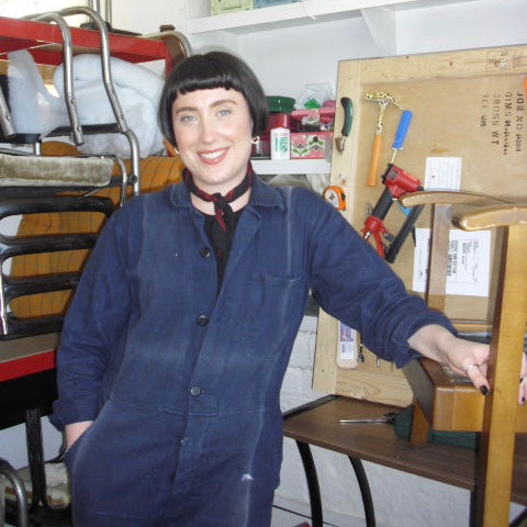 A person with a short black wig stands in a cluttered workshop or studio, wearing a dark blue denim work jacket over a black shirt and red necktie. Shelves and tools are visible in the background.