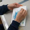 A close-up of a person's hands creating abstract art on a white rectangular canvas with blue brushstrokes and yellow thread, set on a gray worktable with tools.
