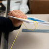 A close-up of a person's hand holding a stack of printed cards or pages bound with a yellow elastic string, with a desk and blue storage box in the background.