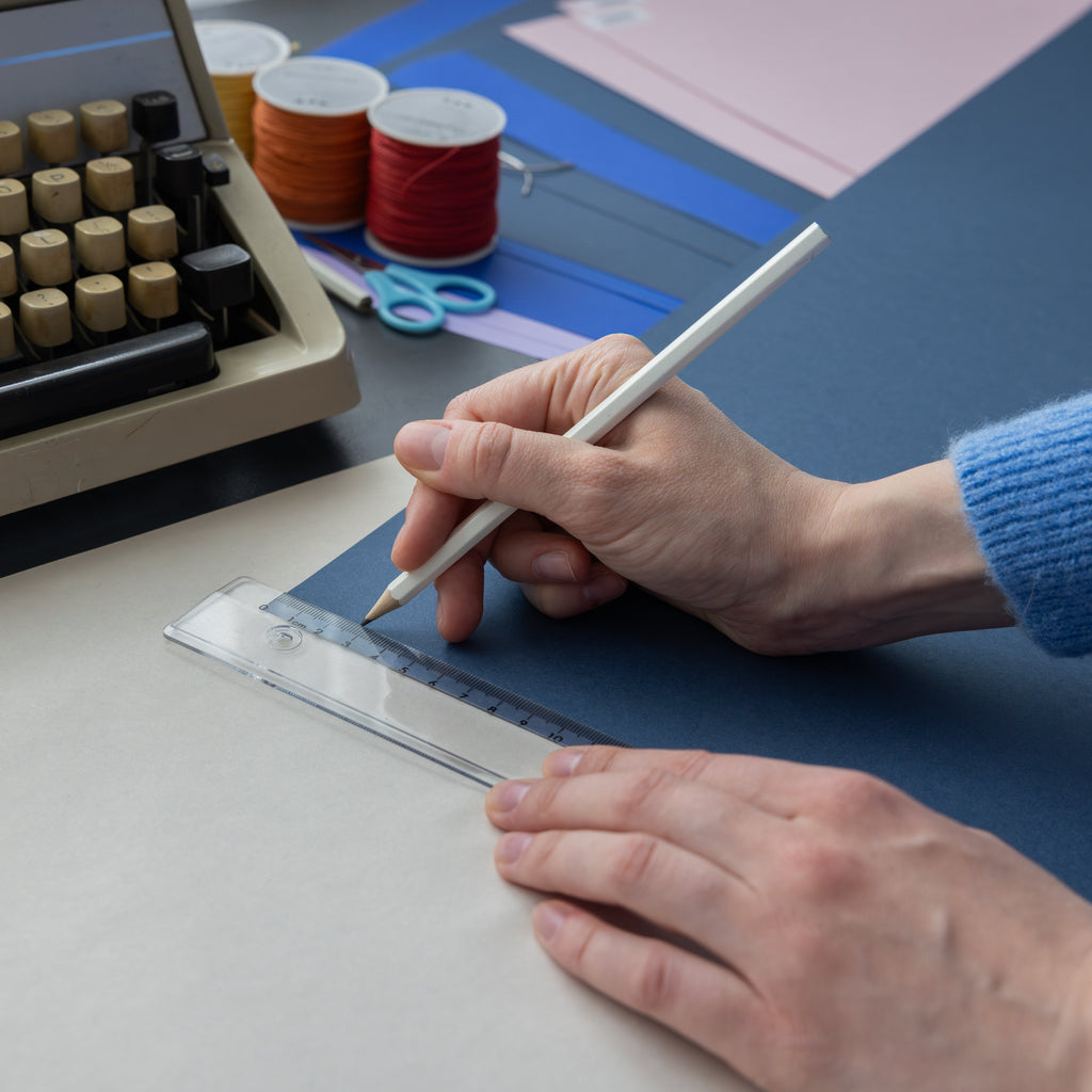 A close-up of a person's hands drawing on a sheet of dark blue cardstock using a white pencil and clear ruler, with a vintage typewriter, craft scissors, and colored thread spools in the background.