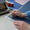 A close-up of a person's hands drawing on a sheet of dark blue cardstock using a white pencil and clear ruler, with a vintage typewriter, craft scissors, and colored thread spools in the background.