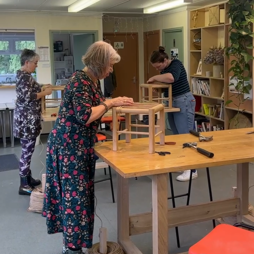 A group of people working together in a bright workshop or classroom, assembling wooden stools at long wooden tables. One person in a floral dress is in the foreground, while others are seen in the background near shelves and tools.