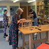 A group of people working together in a bright workshop or classroom, assembling wooden stools at long wooden tables. One person in a floral dress is in the foreground, while others are seen in the background near shelves and tools.