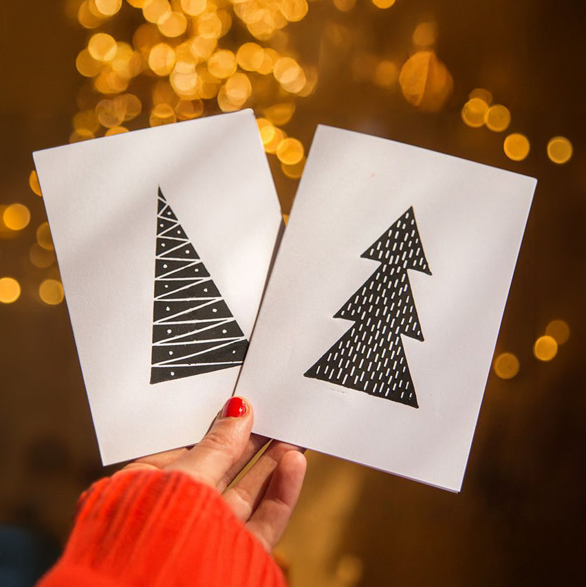 A hand wearing a bright red knit sweater holds two white square cards featuring minimalist black Christmas tree designs, set against a warm, out-of-focus background of golden bokeh lights.