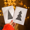 A hand wearing a bright red knit sweater holds two white square cards featuring minimalist black Christmas tree designs, set against a warm, out-of-focus background of golden bokeh lights.