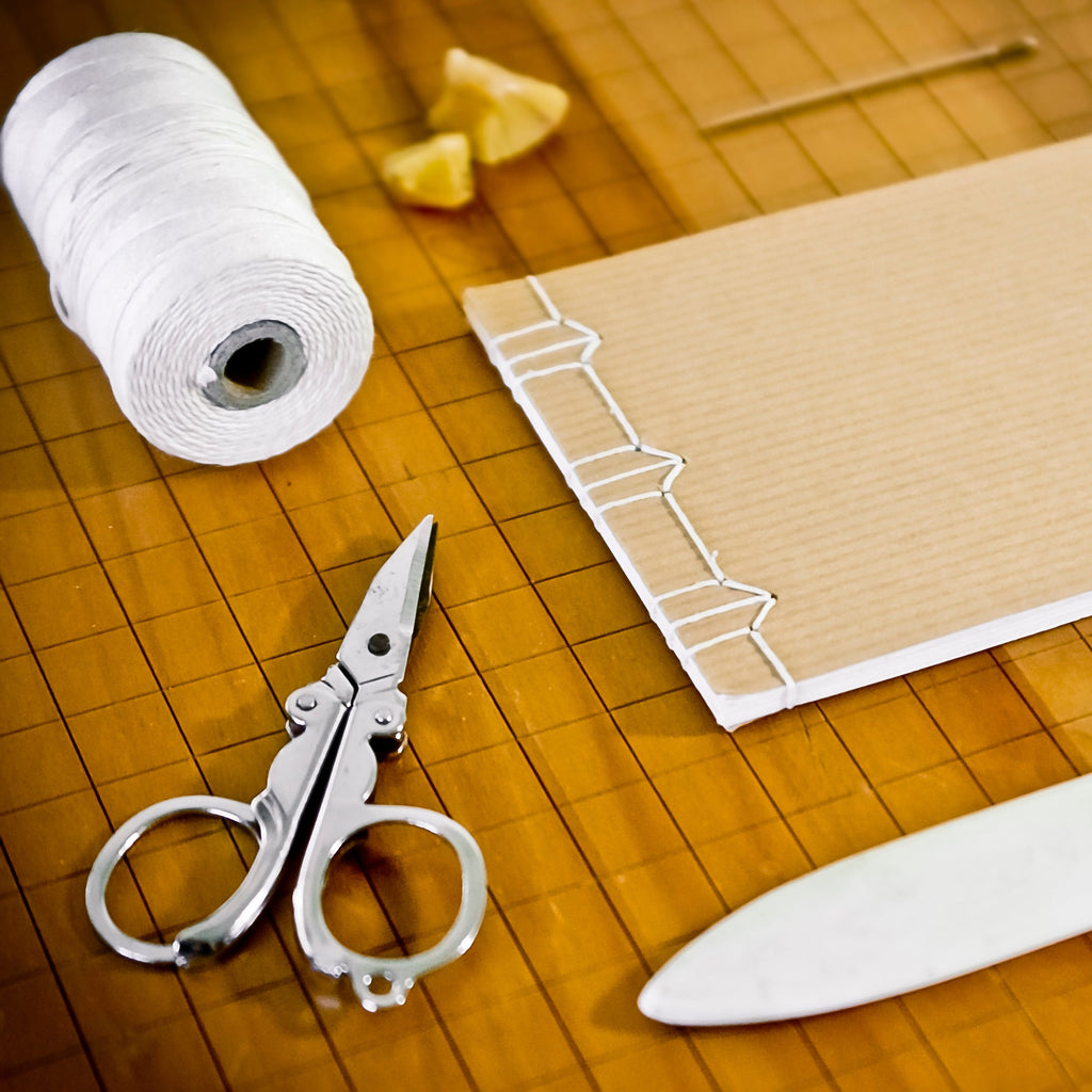 A close-up of a handmade bookbinding or notebook-making setup on a wooden surface, including a spool of white thread, a small notebook with wire binding, scissors, a wooden tool, and a white leather blank.