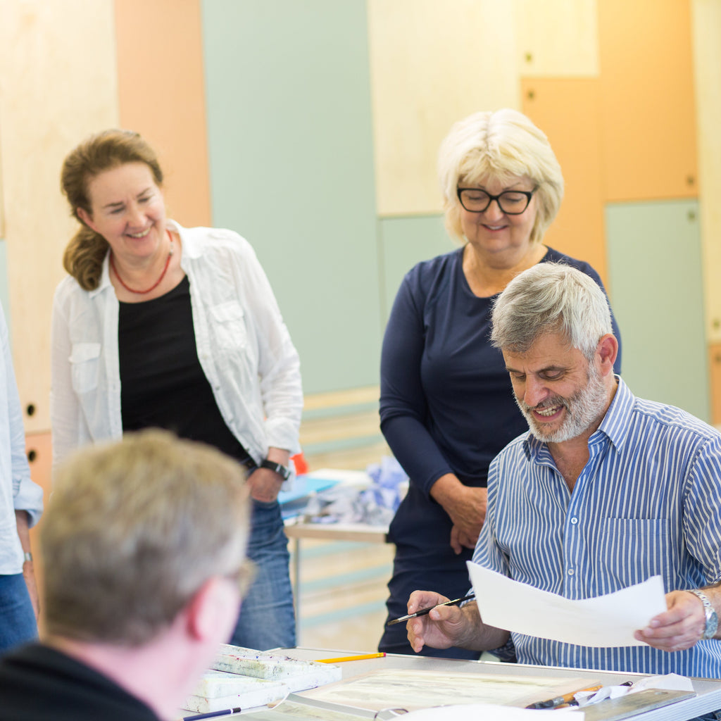 A group of six people gathered around a table in an indoor office or meeting space, with one man in a blue striped shirt holding and reading a document while others observe.